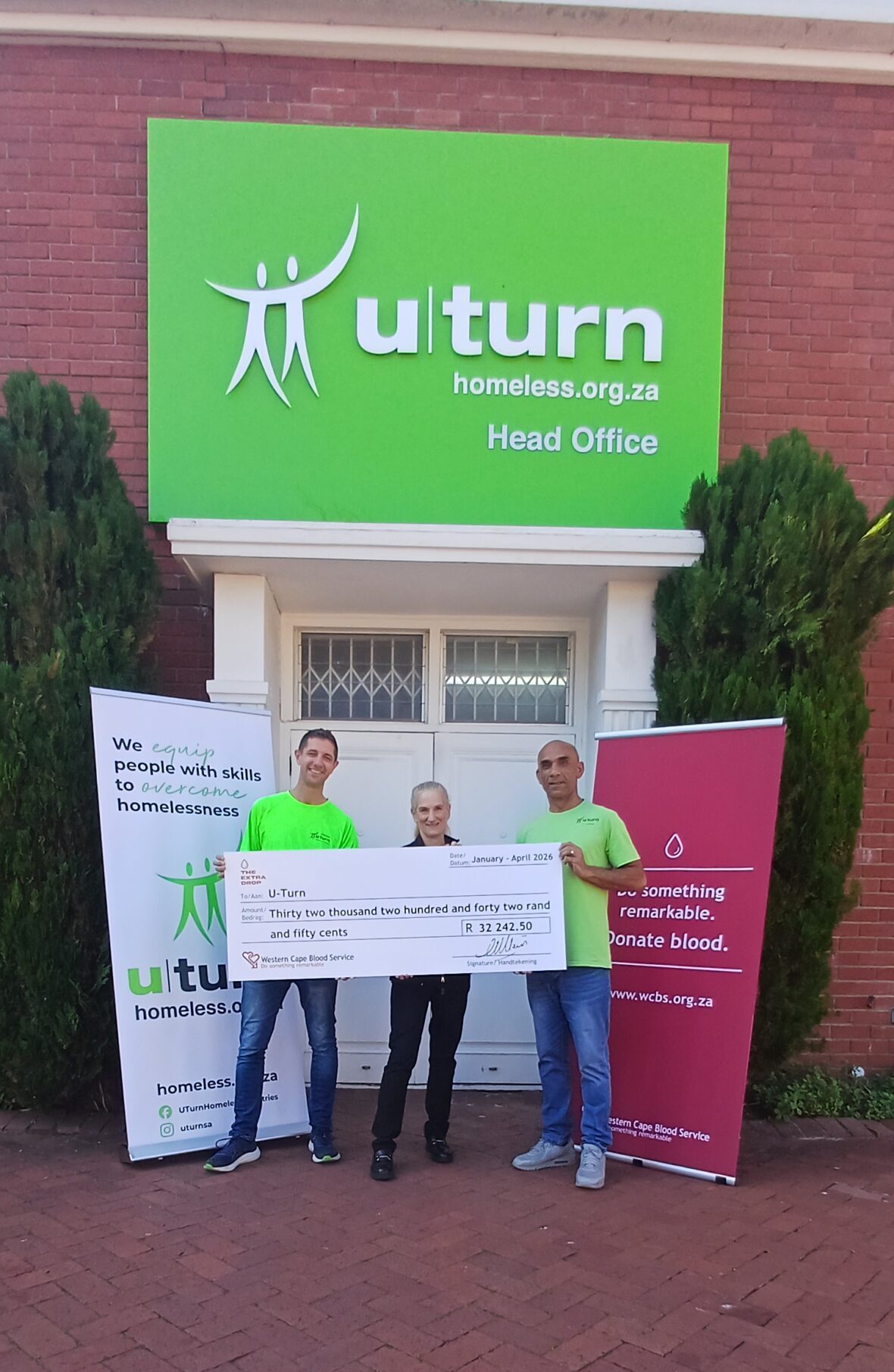 20260520_114933 Three people in neon green shirts hold a large ceremonial cheque outside a brick building with a bright green U-turn sign above them.