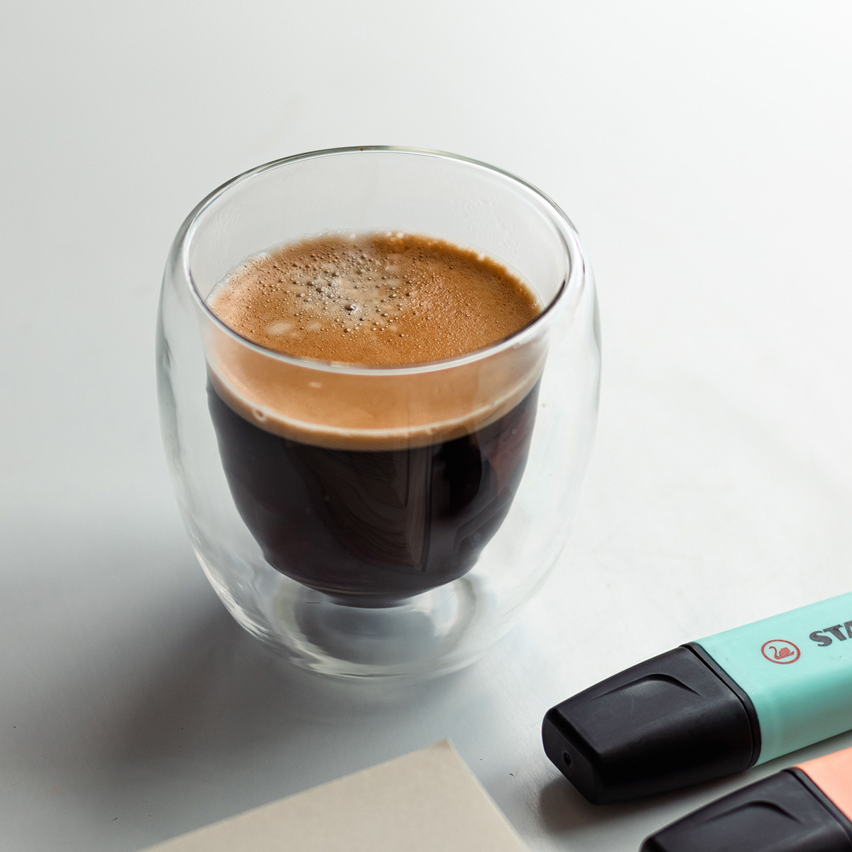 Double-walled glass cup of black coffee with crema on a white surface, pastel highlighters nearby.