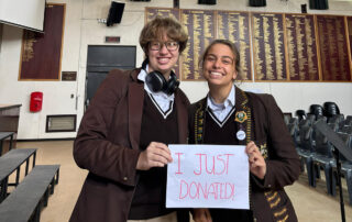 Two smiling students in brown school uniforms hold a handwritten sign that reads 'I JUST DONATED!' inside a classroom or assembly hall.