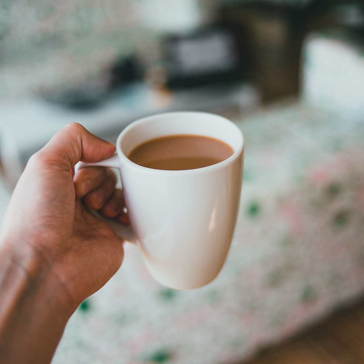 Close-up of a hand gripping a white mug filled with hot coffee, against a softly blurred home interior background.